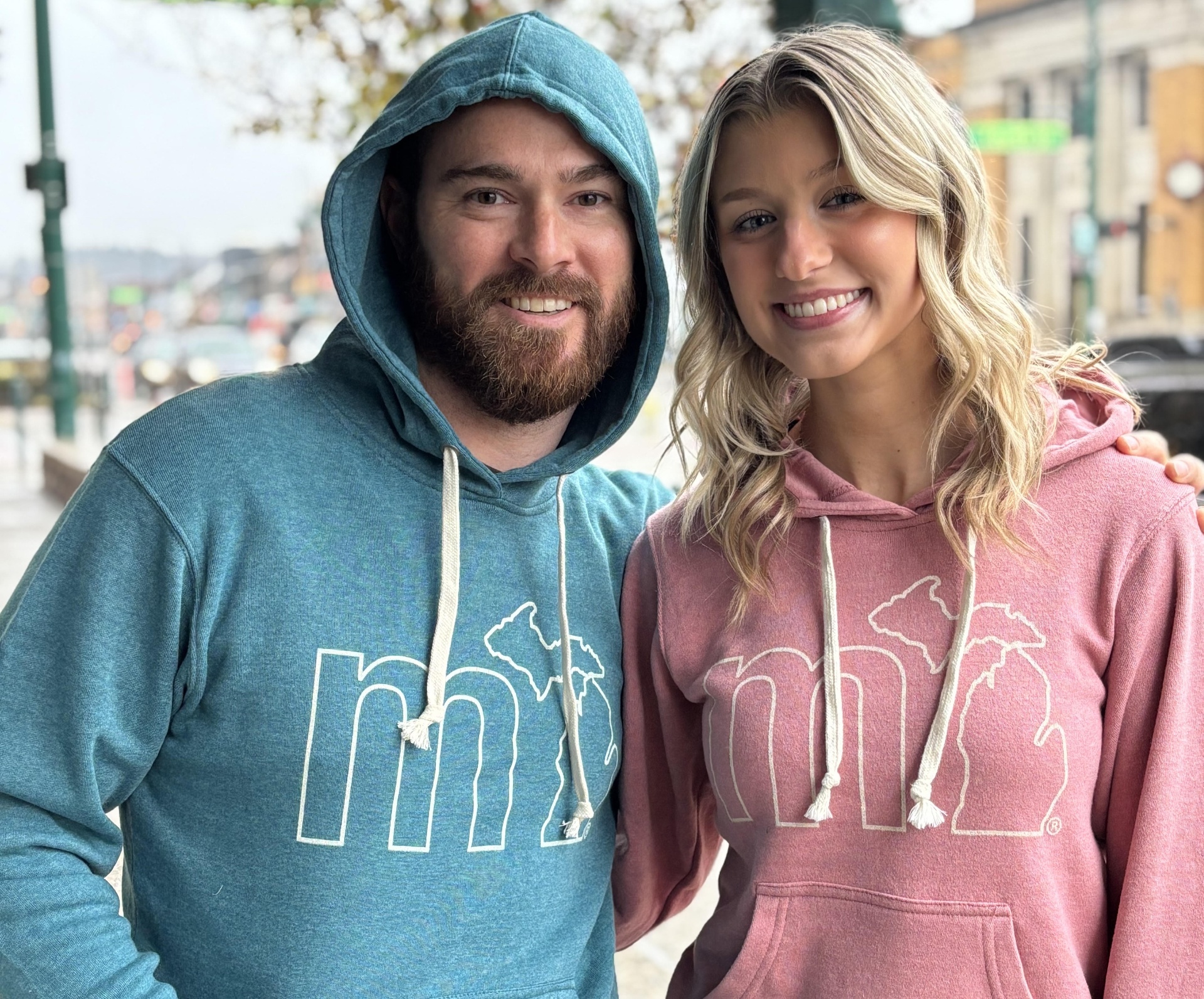 Man and woman wearing teal and rose-pink MI hoodies on a sunny downtown street